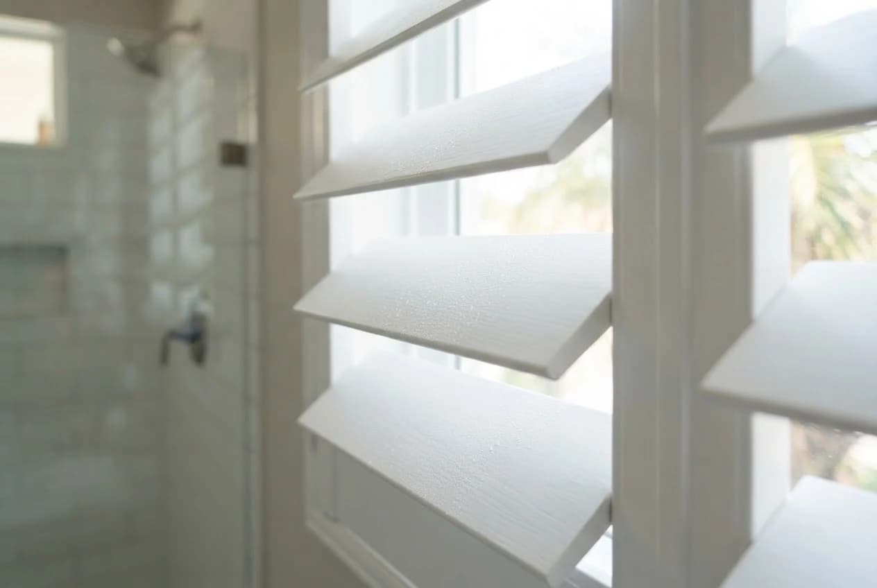 Close-up of white waterproof faux wood plantation shutters with moisture droplets on the louvers beside a glass walk-in shower.